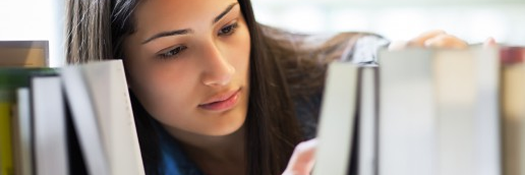 A photo of a young woman with long dark hair looking at books on a bookshelf. She is reaching for a book with her right hand and is looking down intently at the books in front of her.